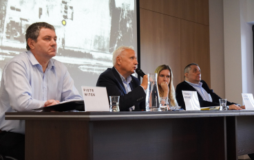 Cztery osoby, trzech mężczyzn i kobieta siedzą przy stole, jeden starszy mężczyzna przemawia do mikrofonu.