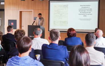 Mężczyzna w beżowej marynarce stoi przy mównicy i przemawia do mikrofonu, w tle prezentacja.