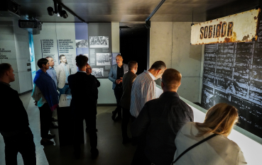 Grupa osób na wystawie, widoczna stara tablica z napisem SOBIBOR.