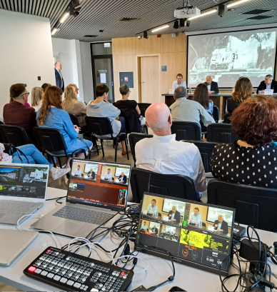 Grupa osób w sali edukacyjnej słuchających debaty, na pierwszym planie laptopy oraz sprzęt elektroniczny służący do transmisji.