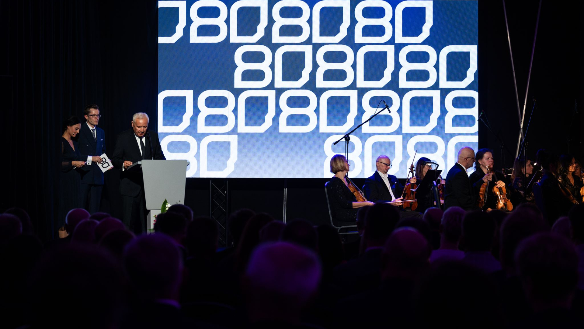 Grupa osób na scenie, prowadzący spotkanie, dyrektor oraz grupa muzyków, w tle ekran z liczbą 80. 