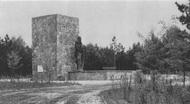 A black-and-white photograph showing a monument at the Sobibór extermination camp depicting a Mother and Child, set on a concrete plinth; in the background, a concrete and stone prism, surrounded by small trees.