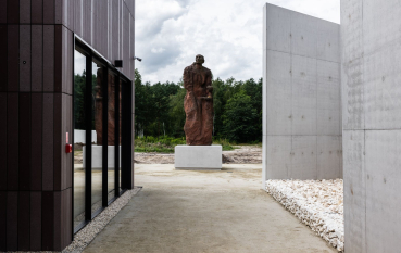 A statue of a mother and child on a concrete basis; on the left, a museum building with large windows; on the right, a section of a concrete wall; the Wall of Remembrance