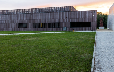 Brown museum building with rectangular, elongated windows; on the right, a statue of a mother cuddling her child and a section of a concrete wall, the Wall of Remembrance; in the background, the setting sun.