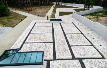 Aerial view showing the remnants of the gas chamber layout covered with white aggregate; on the left, a glass panel revealing the original foundations