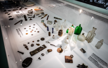 Items: small metal buttons, coloured glass bottles, toothbrushes, coins in a milky-white glass display case.
