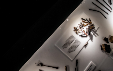 A section of the illuminated display case; items on display: a man’s shoe, a knife, a fork, bullets and long nails.