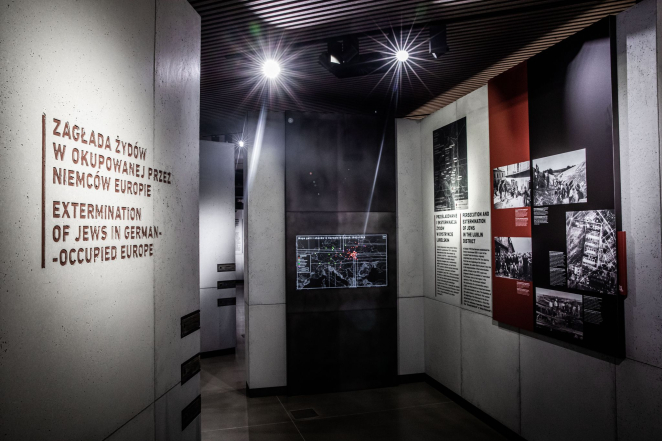 View of the exhibition, a concrete wall visible on the left with the engraved words: extermination of Jews in German-occupied Europe; on the right information panels with documents and archival photographs.