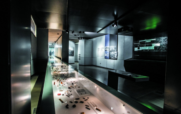 A view of the permanent exhibition, with an illuminated display case containing artefacts in the foreground, historical exhibition panels in the background, and a green light visible. 