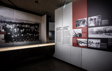 A view of the exhibition: on the left, a large black-and-white photograph on the wall depicting a large group of men; on the right, a panel with historical information and photographs, with the colour red dominating the panel.