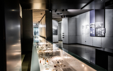 A view of the exhibition: a milky-white, illuminated glass display case containing objects; to the right, panels featuring historical information and photographs; the dominant colour of the panels is purple.