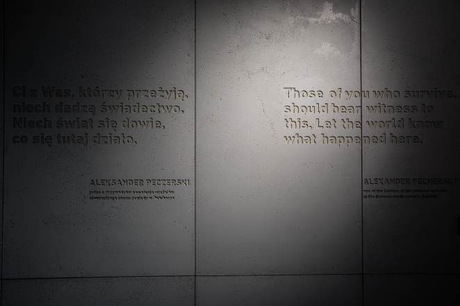 An engraved concrete wall bearing an inscription in Polish and English, half of which is obscured by shadow; the lettering is blurred.