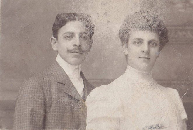 A sepia-toned photograph showing a middle-aged couple: on the left, a man with a moustache and dark hair combed to the sides with a prominent parting; on the right, a woman in a white lace dress with a stand-up collar and her dark hair pinned up high.