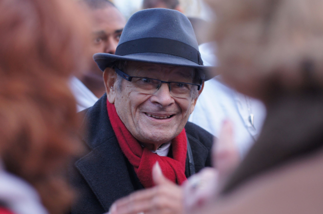 Elderly smiling man wearing a hat, glasses, and a red scarf.