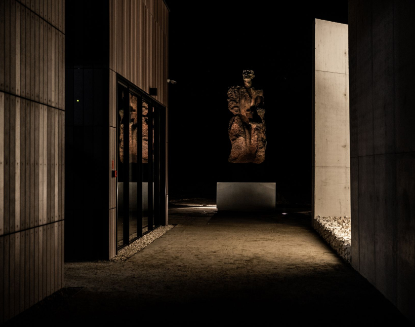 A night-time photograph showing the entrances to the Museum and Memorial in Sobibór; on the right, a glass-panelled wall; in the centre, a monument to a Mother and Child; on the right: a section of concrete wall – the Wall of Remembrance.