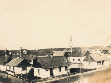 Kilka domów otoczonych żywopłotem, po lewej stronie tablica: SS Sonderkommando i dwie nazistowskie flagi.