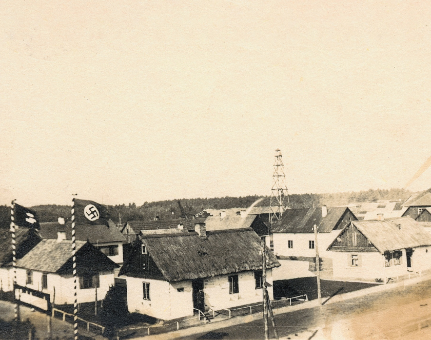 Kilka domów otoczonych żywopłotem, po lewej stronie tablica: SS Sonderkommando i dwie nazistowskie flagi.