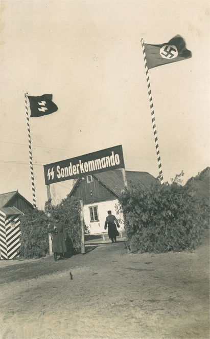 Biały budynek ze spadzistym dachem, brama z napisem SS-Sonderkommando, po obu stronach nazistowskie flagi, w centrum mężczyzna ubrany w niemiecki mundur.