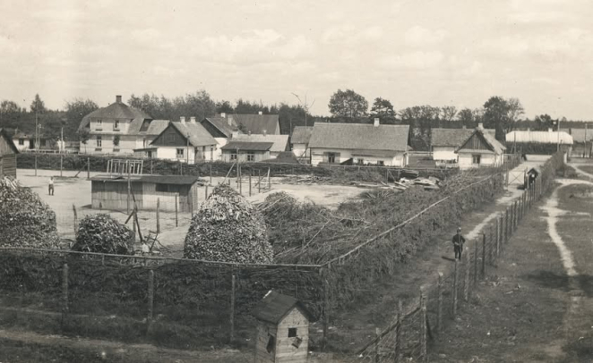 Liczna zabudowa niskich białych domów, podwójne ogrodzenie z drutu kolczastego i gałęzi.