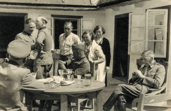 Grupa osób, mężczyzn w niemieckich mundurach  oraz kobiet w cywilnych przy stole nakrytym kieliszkami oraz szklankami, w tle biały dom. 