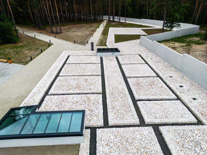 Fundamenty komór gazowych pokryte białym kruszywem, po lewej przeszklone okno czasu prezentujące wygląd archeologicznie zachowanych fundamentów