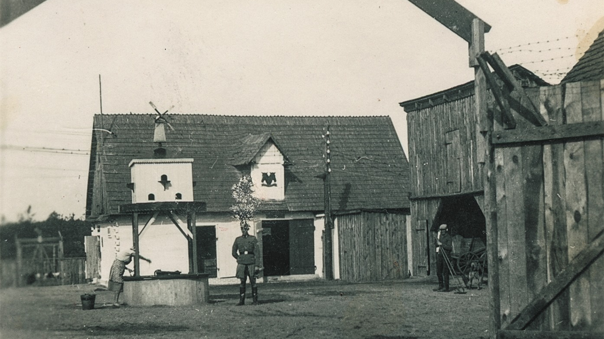 Czarno-białe zdjęcie przedstawiające otwartą przestrzeń ze studnią i białym, parterowym budynkiem w tle. Przy studni stoi mężczyzna ubrany w niemiecki mundur. Na pierwszym planie widoczna brama, nad którą znajduje się ozdobny napis Erbhof.