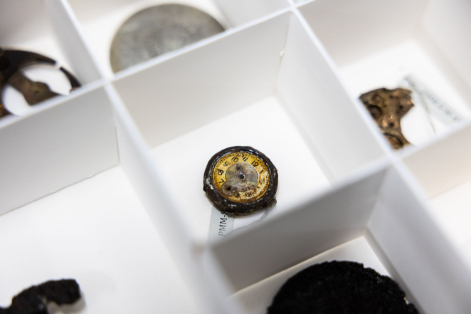 Metalowe drobne przedmioty w bezkwasowych pojemnikach z separatorami, w centrum mała tarcza zegarka bez wskazówek. 