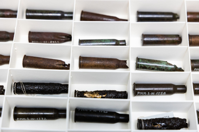 Kilkanaście łusek pocisków w bezkwasowych pojemnikach z separatorami, na części z nich widoczny numer inwentarzowy. 