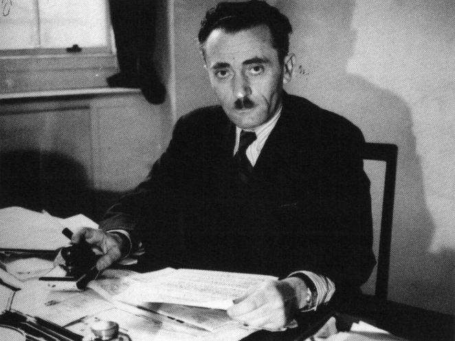 Czarno-białe zdjęcie: mężczyzna w średnim wieku o czarnych włosach i prostymi wąsami typu szczoteczkowego trzyma w rękach dokument.