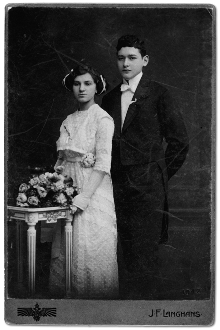 Czarno-białe zdjęcie ślubne:  kobieta ubrana w białą koronkową suknię ślubną  o upiętych wysoko czarnych włosach, mężczyzna ubrany w czarny garnitur, białą koszulę z biała muszką; przed parą młodą stolik i ułożony na nim bukiet jasnych kwiatów.  