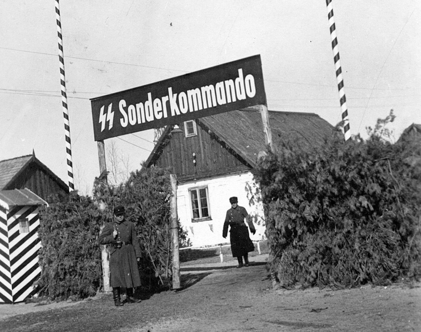 Czarno-białe zdjęcie. Wejście do ogrodzonego obszaru z tablicą „SS Sonderkommando”. Przy bramie stoją dwaj umundurowani strażnicy, a w tle widoczny jest drewniany budynek.