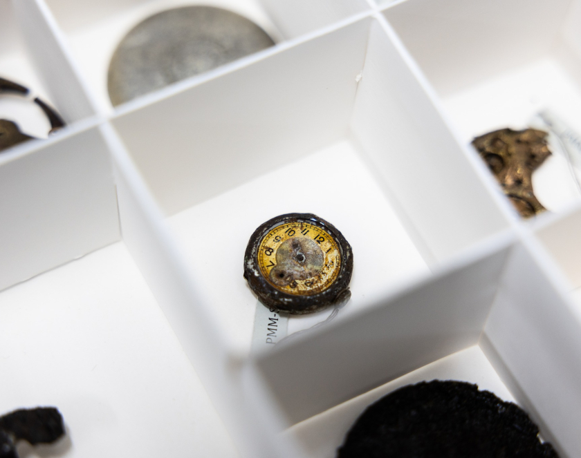 Close-up of a rusted dial from a small watch or pocket watch with a yellow face and visible numbers, placed in a white compartment of a museum display case. Other small metal objects and fragments of jewelry are visible in the neighboring compartments.