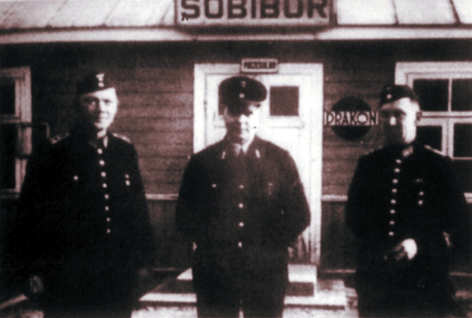 Czarno-białe zdjęcie przedstawiające trzech mężczyzn ubranych w czarne mundury kolejarzy stojących przed drewnianym, parterowym budynkiem. W tle widoczne białe drzwi wejściowe, nad którymi wisi duża, prostokątna, biała tablica z czarnym napisem Sobibór.