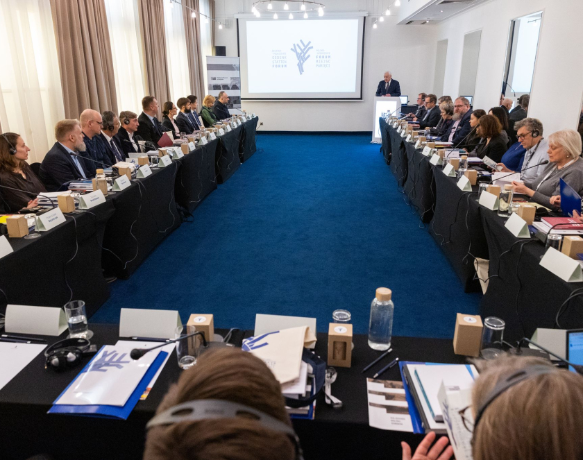 Ludzie elegancko ubrani w sali konferencyjnej siedzący przy stołach ułożonych w podkowę, jedna osoba przy mównicy, na ścianie wyświetlany baner.   