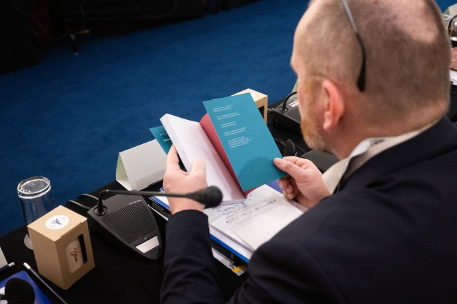 Mężczyzna trzyma w ręku książkę, na stole mikrofon oraz materiały biurowe. 