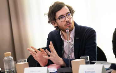 Mężczyzna w okularach z dłuższymi włosami ubrany w garnitur przemawia do mikrofonu.